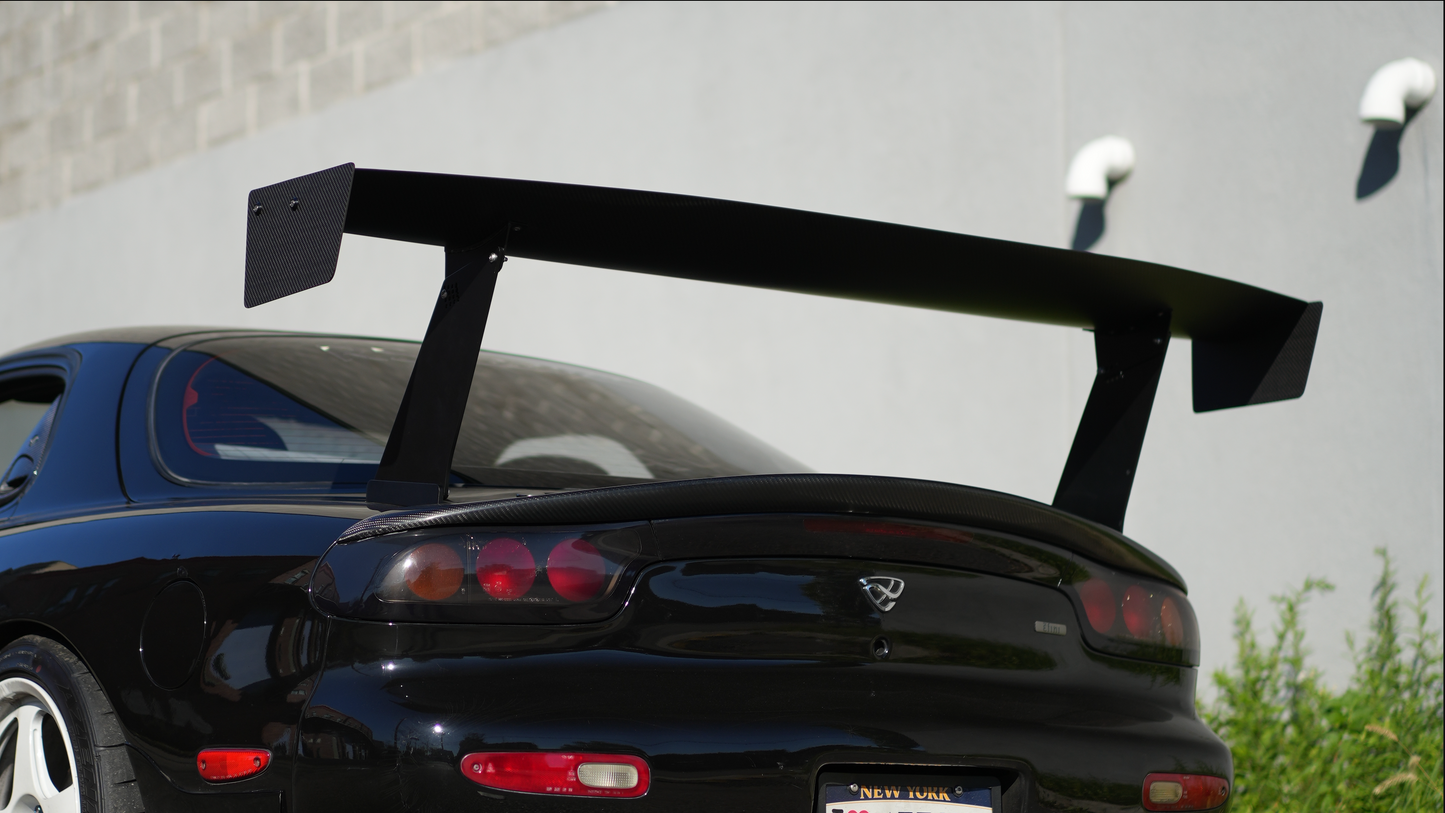 A black sports car shown from the rear, featuring a large black rear wing spoiler mounted above the trunk, parked outdoors against a light gray wall.