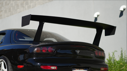 A black sports car shown from the rear, featuring a large black rear wing spoiler mounted above the trunk, parked outdoors against a light gray wall.